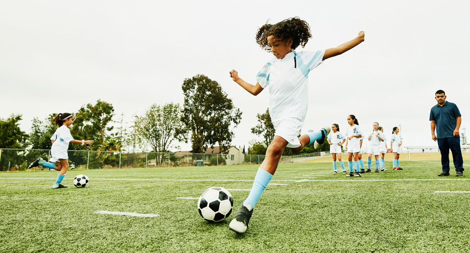 A young girl kicking a football, wearing white and blue kit. Her team and coach are in the background.